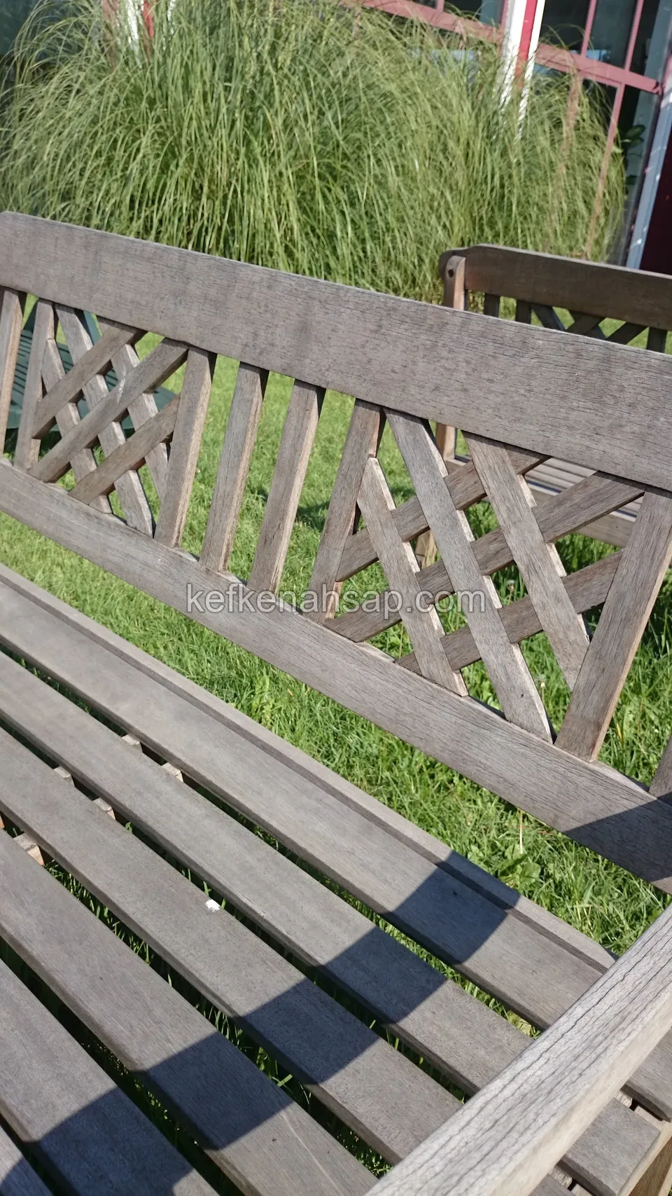Nahansicht von Gitterlehne und Lattensitz einer sonnengrauen Holzgartenbank, auf dem Gras liegen Lattenschatten