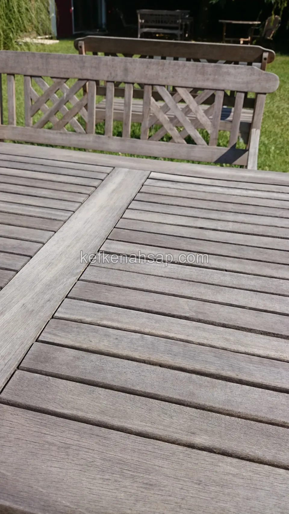 Von der Sonne ergraute Lattenplatte eines Holztisches mit der Lehne einer Bank dahinter