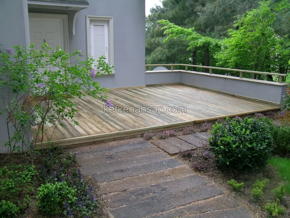 Gri boyalı evin önünde, alçak duvarla sınırlanan gri tonlu deck; önünde traverslerden döşenmiş bir yürüyüş yolu