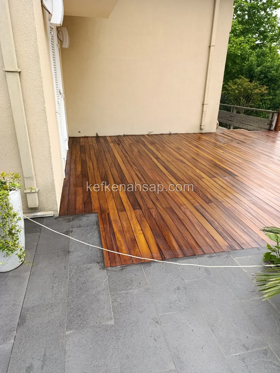 Glossy red-brown deck laid beside grey stone flooring at the foot of a building wall, with a thin string line on the floor