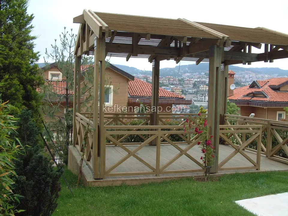 Çimlik bahçede çapraz korkuluklu ahşap kamelya, çıtalı çatı, korkuluğa sarılmış gül ve arkada kiremit çatılı evler