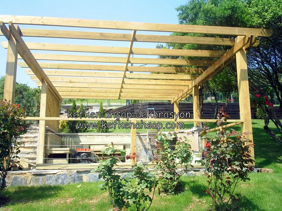 Gül çalıları arasında geniş, çıtalı çatılı açık renk ahşap pergola, payandalı köşe ve arkada kafes panel