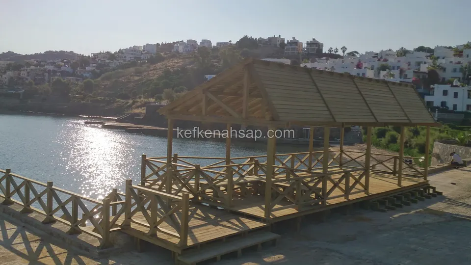 Gün batımında su kenarındaki ahşap kamelya ve çapraz korkuluklu deck, suda ışık yansıması ve arkada yamaç evler