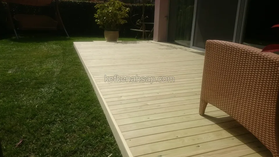 Çimenlik içinde uzanan açık renkli deck zemini, sağda hasır koltuk, arkada cam sürgülü kapı ve saksı