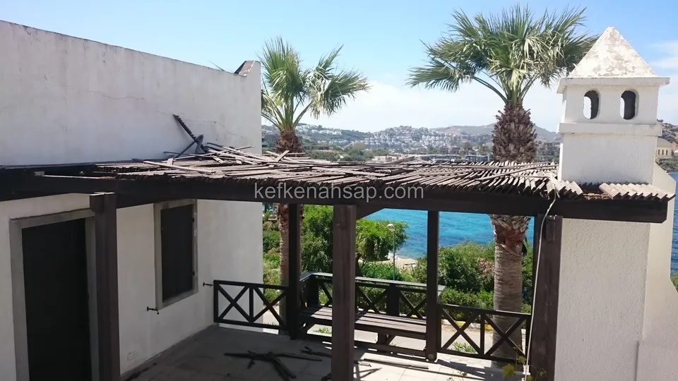 Dunkle Holzpergola mit Schilfabdeckung auf der Terrasse eines weißen Hauses, Geländer mit Kreuzstreben, Palmen und Meer