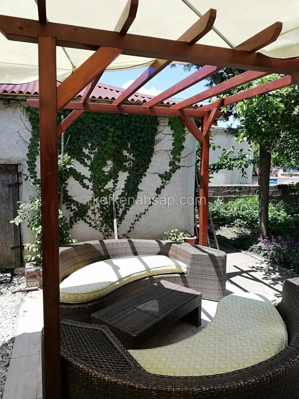 Kırmızımsı vernikli pergolanın altında hasır köşe oturma grubu ve krem tente, arkada sarmaşıklı duvar