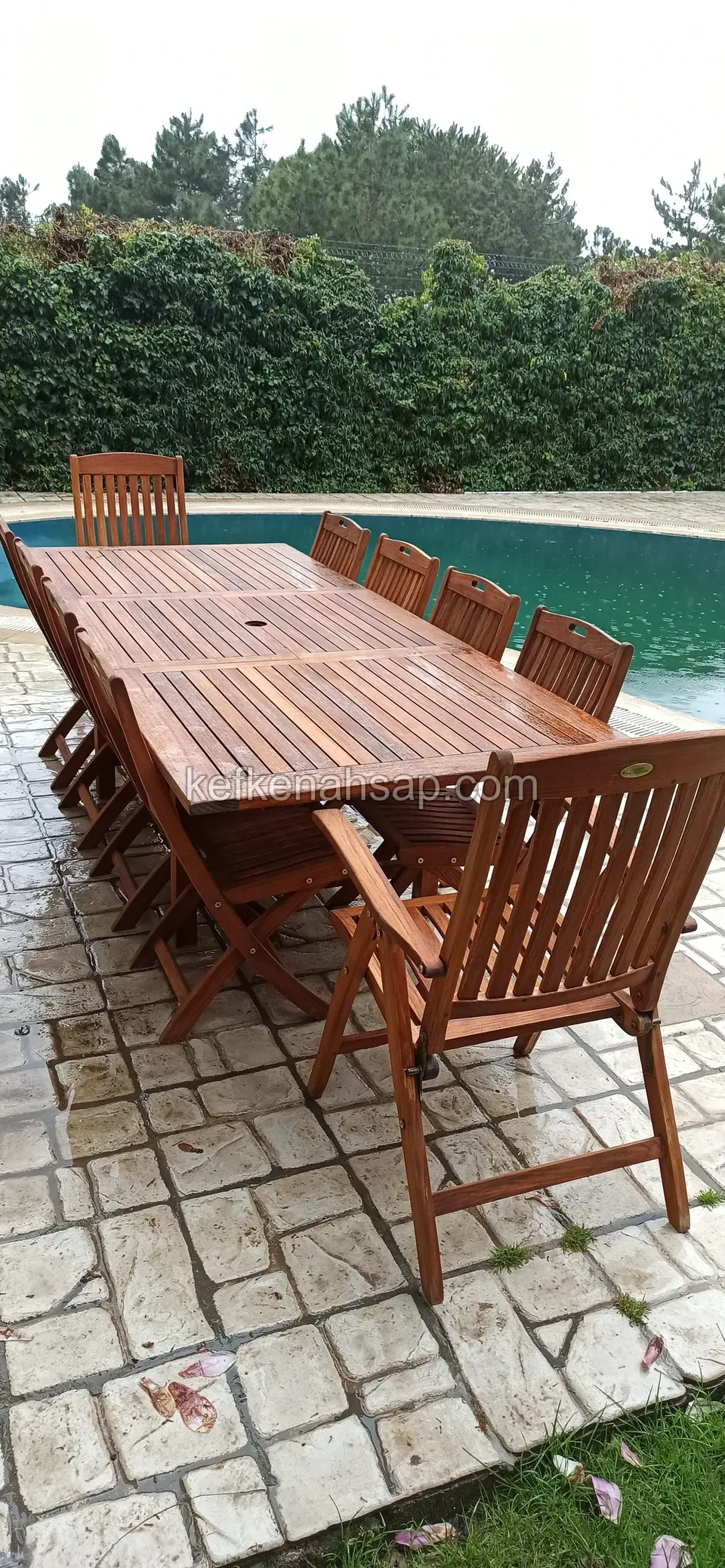Langer Holzesstisch mit Latten, von Klappstühlen umgeben, auf Steinboden am Poolrand