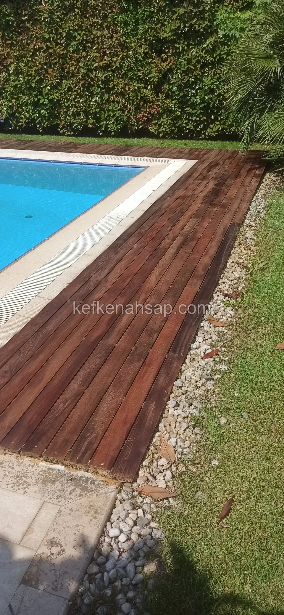 Dark brown deck around a blue pool, separated from the lawn by a strip of white pebbles