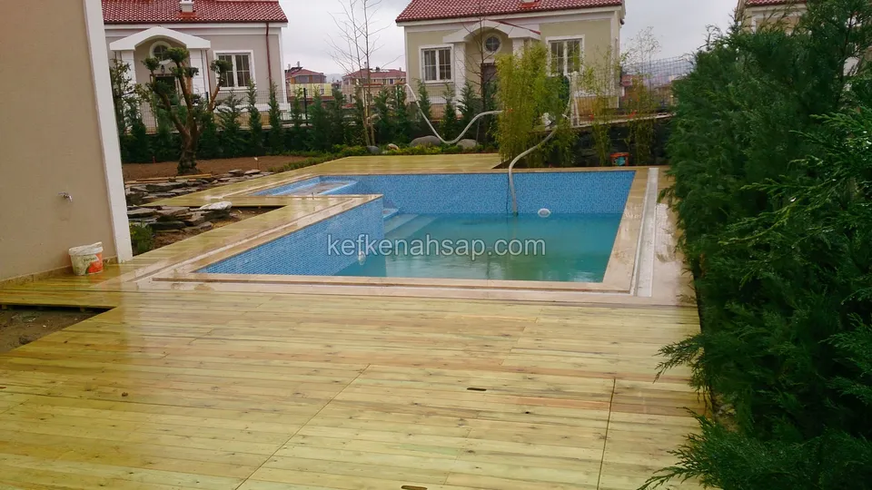 Havuzun çevresini saran geniş açık renkli deck; solda ev duvarı, arkada çalılar ve villalar
