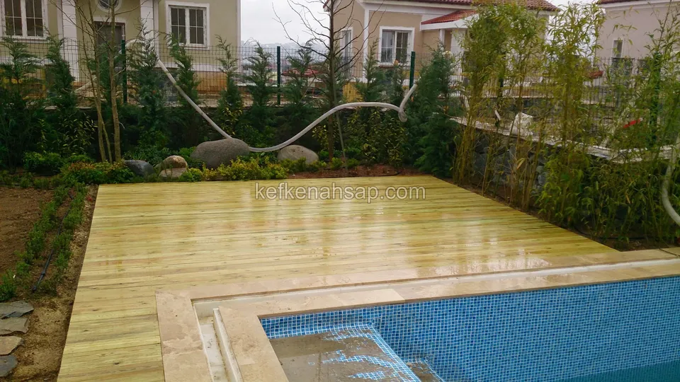 Mozaik fayanslı havuzun çevresine döşenmiş yeni açık renkli deck; arkada çalılık, bambu ve kiremit çatılı villalar