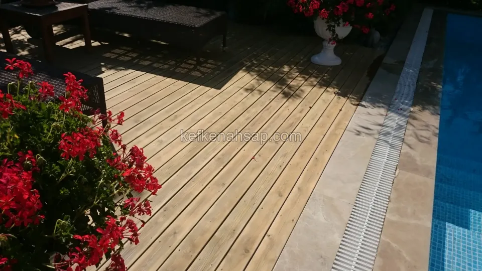Havuz kenarında açık tonlu ahşap deck zemini, yanında taş kenarlık ve kırmızı sardunyalar