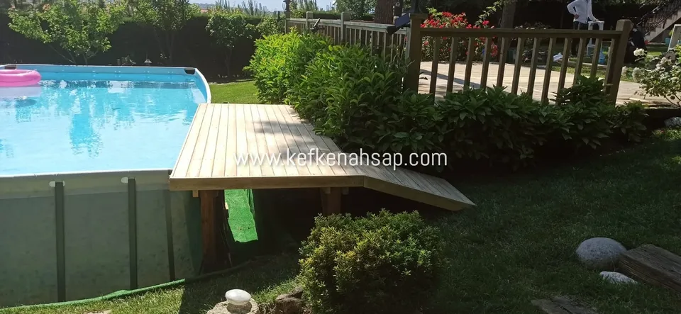 Raised wooden deck platform with a small ramp beside an above-ground garden pool, with a wooden railing behind