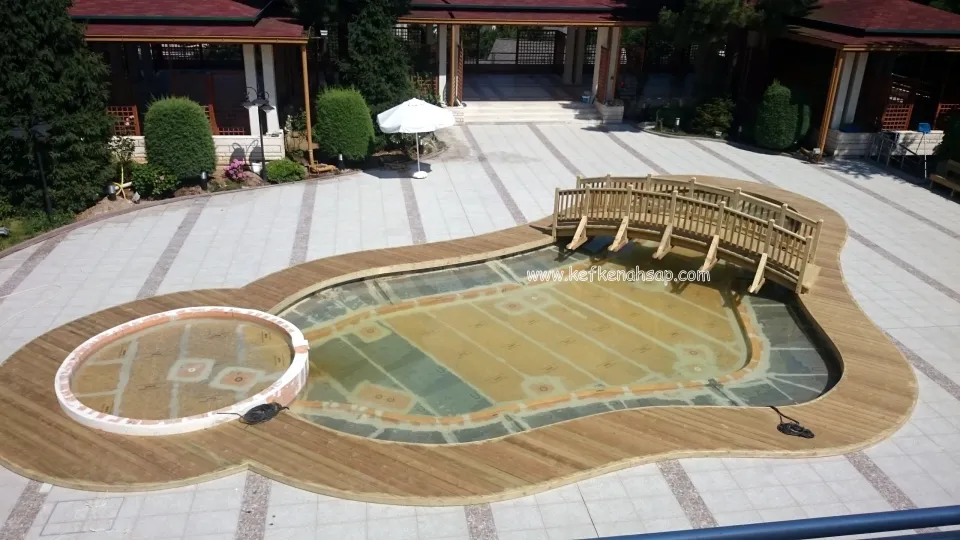 Serbest formlu havuzun çevresinde ahşap deck ve havuzun üzerinden geçen korkuluklu küçük ahşap köprü, arkada bahçe ve köşkler
