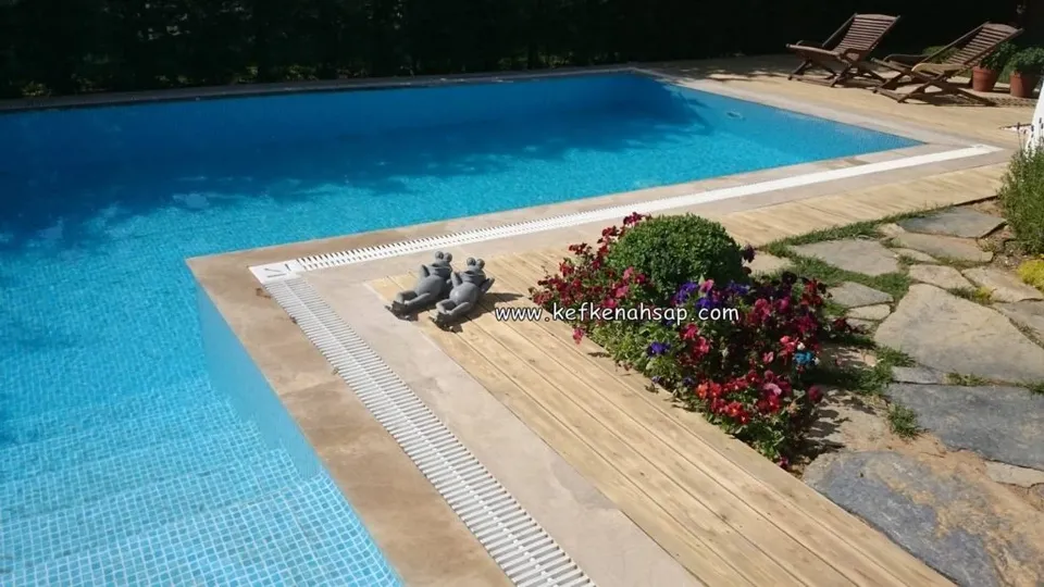 Mavi mozaikli havuzun köşesinde oluklu ahşap deck, yanında çiçek tarhı, doğal taş döşeme ve iki şezlong