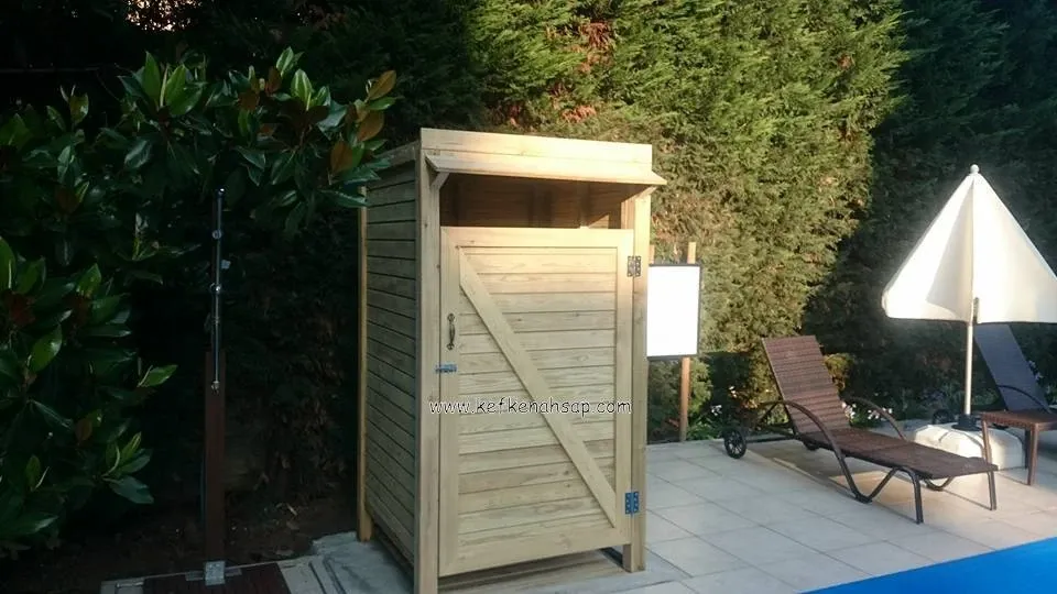 Havuz kenarında, bahçede duran açık renkli ahşap kabin; çapraz destekli kapısı ve üstünde çıkıntılı bir saçak var.