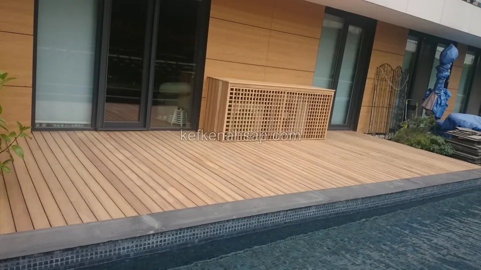 Ahşap kaplı cephenin ve cam kapıların önünde havuza kadar uzanan turuncu deck, duvara yaslı kafesli kapak kutusu