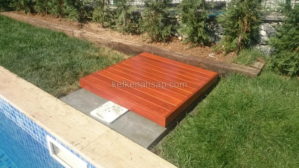 Reddish square timber deck platform on a stone base on the lawn beside a pool, with a cover next to it