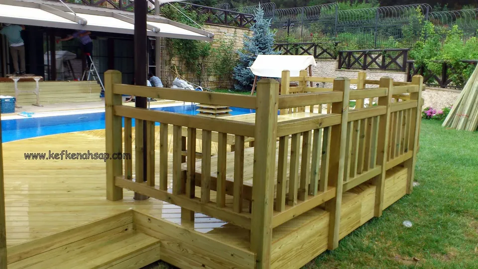 Yeni açık yeşilimsi deck platformunun dikey çıtalı korkuluğu ve köşe dikmesi; arkada havuz, çalışan biri ve bahçe duvarı