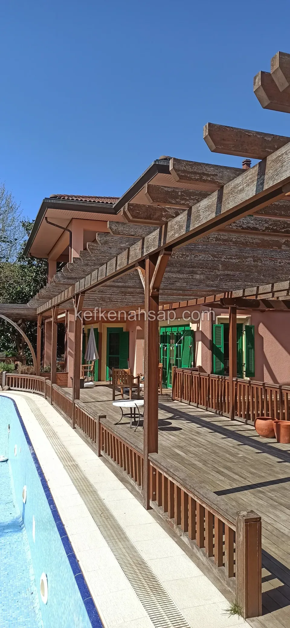 Havuz boyunca uzanan ahşap korkuluklu deck ve üstünü örten koyu tonlu pergola; arkada yeşil panjurlu ev