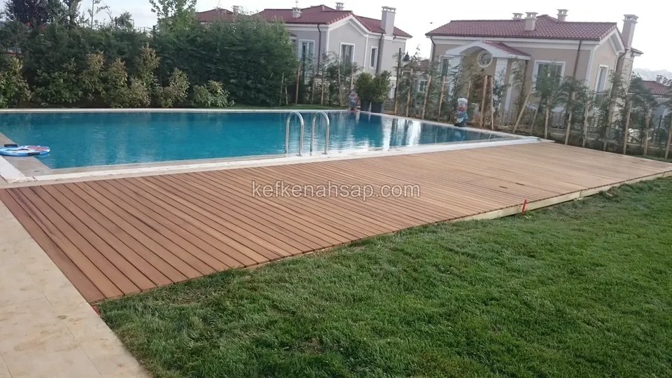 Havuzun kenarında çimle sınırlanan kahverengi deck yüzeyi, arkada kiremit çatılı evler
