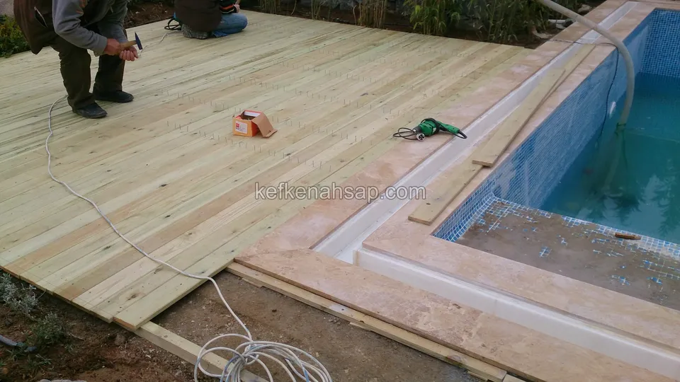 Havuzun taş kenarına kadar döşenmiş açık renkli deck tahtaları; iki usta tahtaları sabitliyor, yerde matkap ve uzatma kablosu