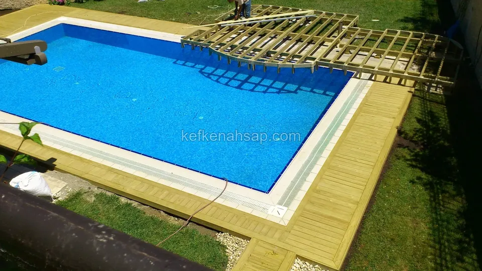 Mavi havuzun kenarını saran açık renkli deck; üst köşede kaplanmamış, kafes gibi duran ahşap iskelet