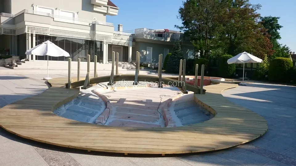 Kavisli bir havuz kabuğunun çevresinde geniş açıdan görünen deck, ön plandaki kavis ve arkada dikmeler, şemsiyeler ve evler