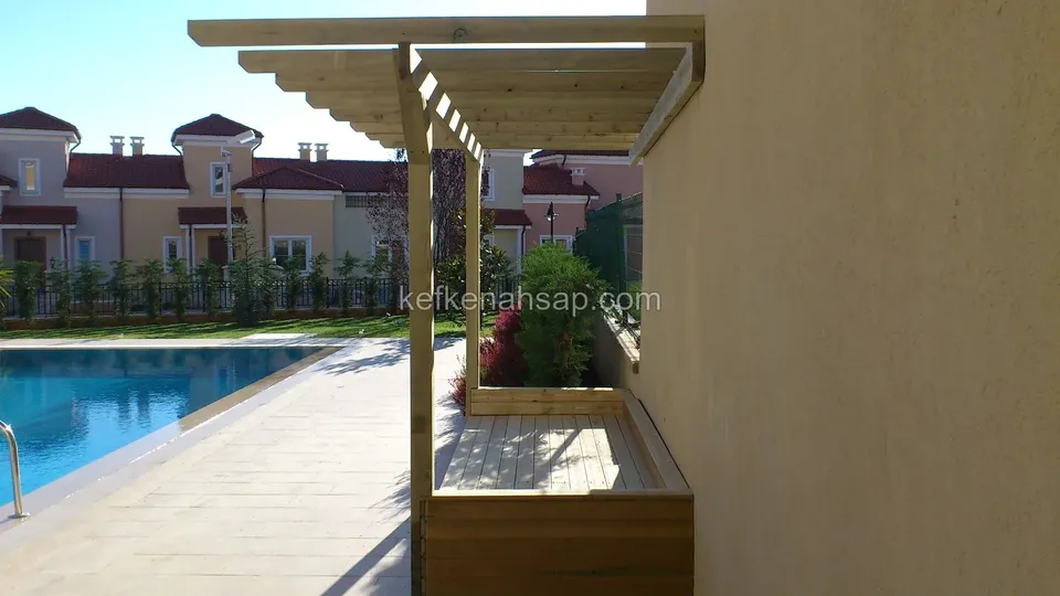 Havuz kenarında duvara yaslanan açık renk ahşap pergola ve altında uzun bitkilik, arkada kiremit çatılı evler