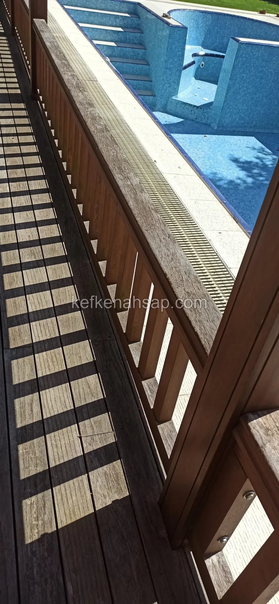 ديكينغ داكن متقادم ودرابزين بني محمر عند حافة المسبح وتسقط ظلال الدرابزين على الألواح