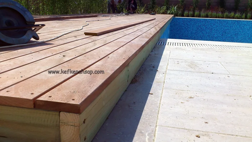 Havuzun ızgaralı kenarında döşenmekte olan kırmızımsı kahverengi deck tahtaları; köşede bir testere