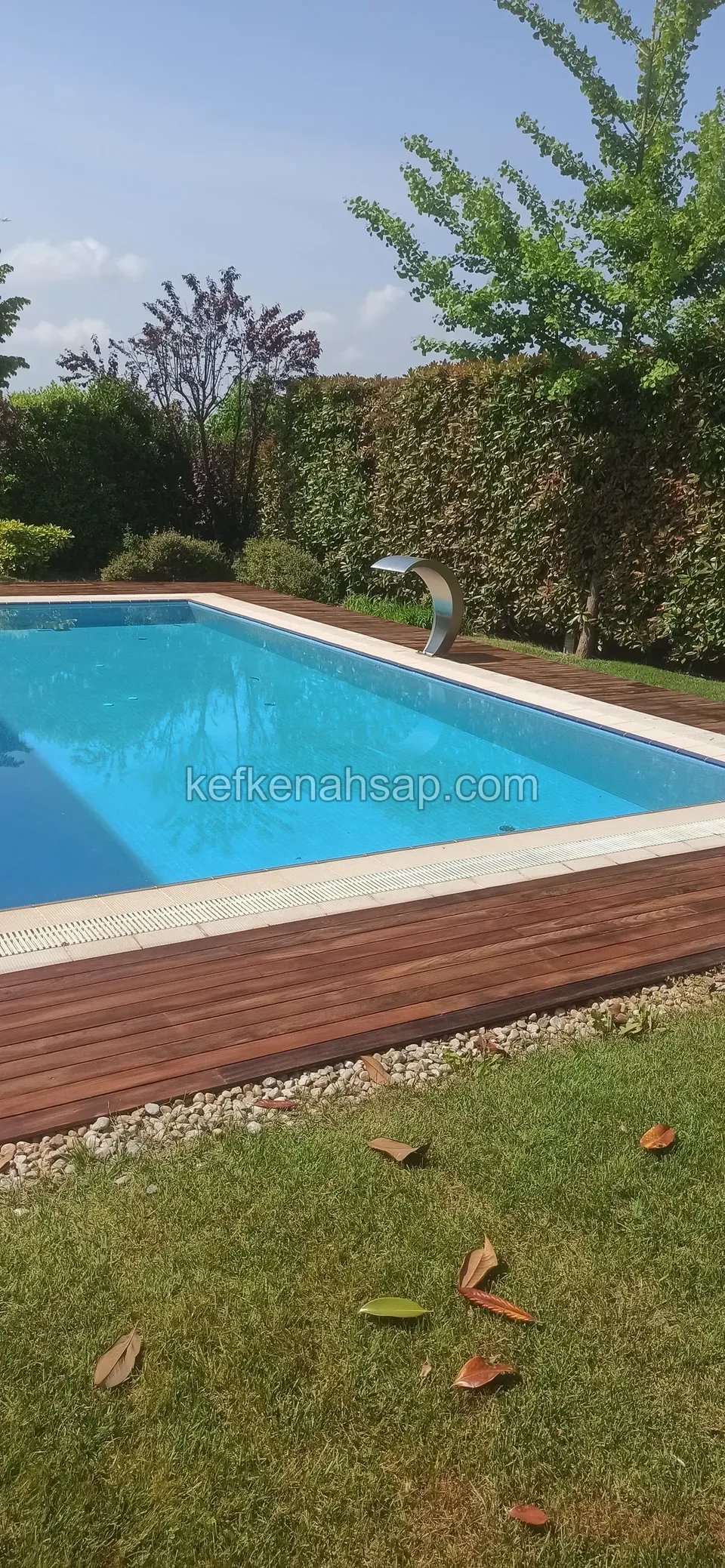 Dark brown deck along the near side of a blue pool, with a pebble strip in front and a trimmed hedge behind