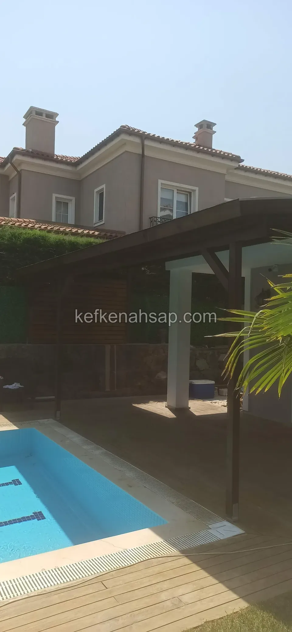 Havuzun kenarında, evin önünde koyu ahşap bir çatı ve ahşap deck; havuzun mavi suyu öne çıkıyor