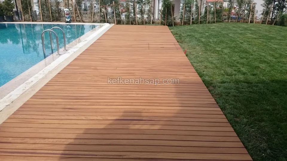 Havuz kenarında turuncu kahve deck şeridi, solda havuz merdiveni, sağda çim ve arkada fidanlar