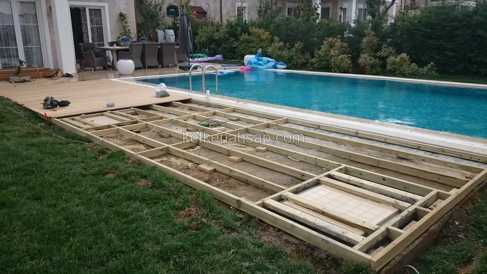 Çimenlik üzerinde geniş ahşap çerçeve, arkada havuz, oturma takımı ve ev; deck tahtaları köşeden başlıyor