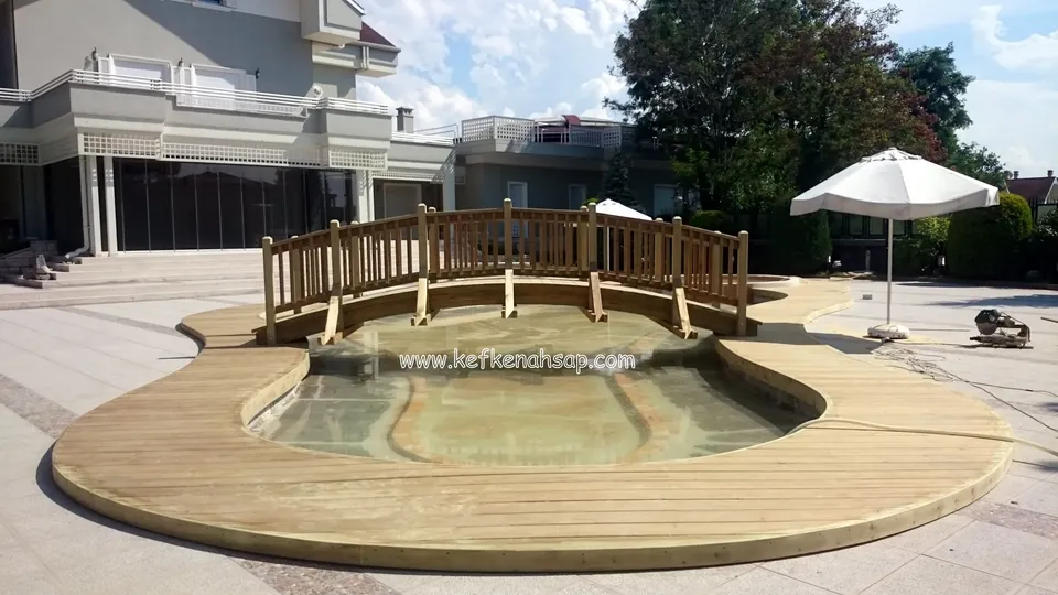 Kıvrımlı ahşap deck ile çevrili havuzun üzerinden geçen kemerli, korkuluklu ahşap köprü