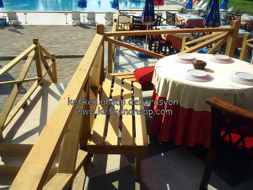 Havuz kenarındaki restoran terasında çapraz çıtalı açık tonlu ahşap korkuluk, ahşap bank ve örtülü yemek masaları