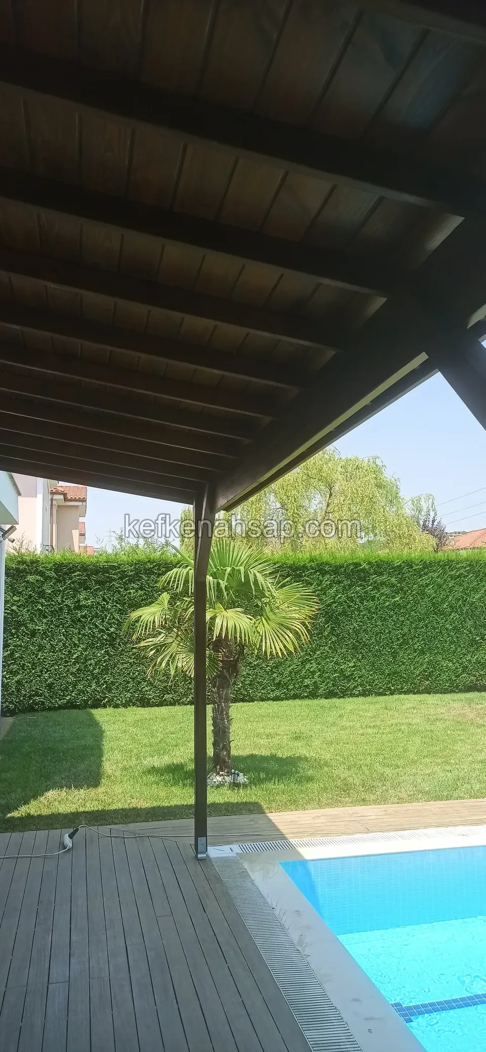 Havuzun kenarında, altında deck zemin bulunan koyu renkli ahşap çatı; arkada çim ve palmiye