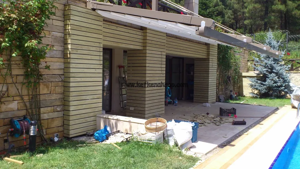 Havuz kenarındaki teras girişinde yatay ahşap tahtalarla kaplanmış kolonlar, taş duvar, çimlik ve yerde aletler, kovalar