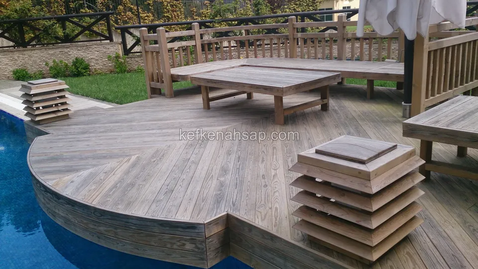Havuzun üzerine doğru kavis çizen, gri tonlu deck platform; üzerinde bank, alçak masa ve kademeli ahşap fenerler