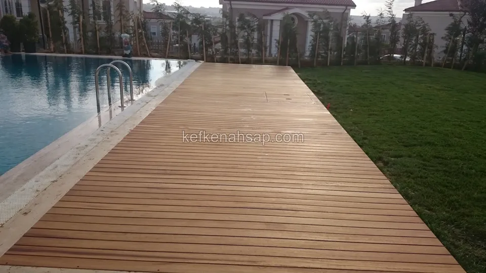 Havuz merdiveninin yanından çimlere doğru daralan uzun deck yüzeyi, arkada ağaçlar ve evler