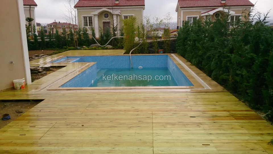 Mavi havuzun çevresinde L biçiminde dönen geniş açık renkli deck; arkada servi sırası ve kiremit çatılı villalar