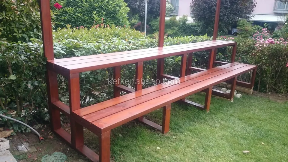 Lange zweistufige Holzbank in Rotbraun vor einem Zaun im Rasengarten