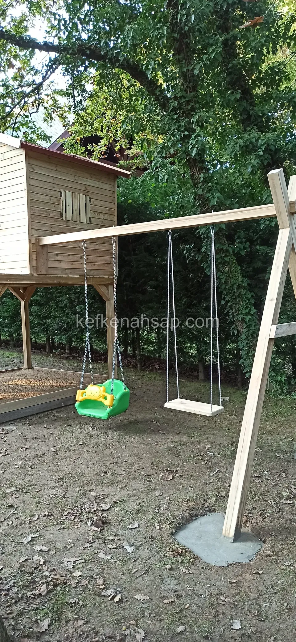 Beton ayağa oturan ahşap salıncakta biri yeşil plastik, biri tahta iki oturak asılı; arkada yüksek zeminli oyun evi