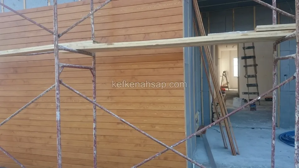 İskelenin arkasında turuncu tonlu yatay ahşap kaplama ve sağda kapı boşluğundan görünen boş iç mekân ile merdiven