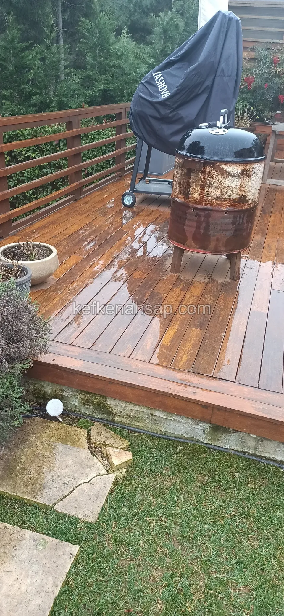 Islak, cilalı deck üzerinde mangal, paslı bir ocak ve ahşap korkuluk; kenarda taş zemin