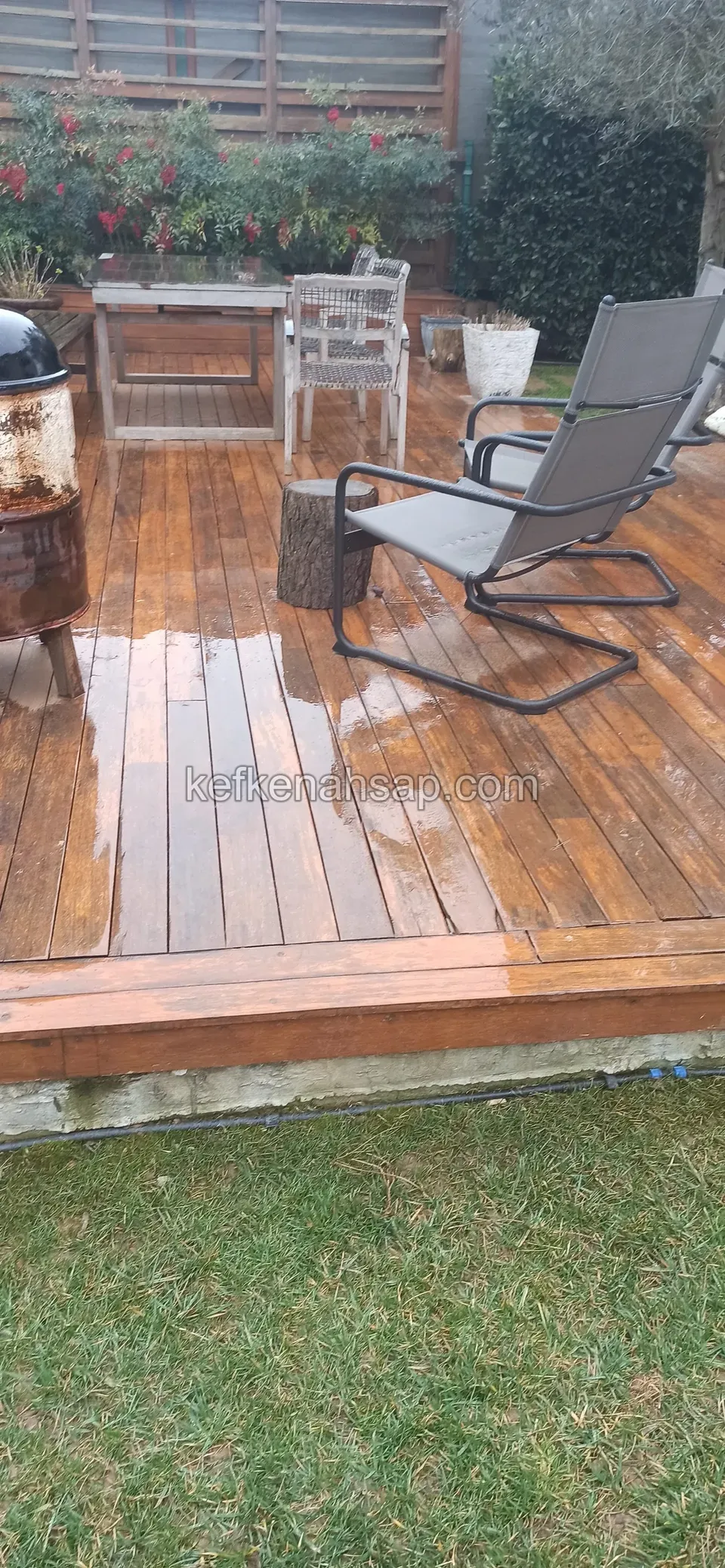 Islak cilalı deck üzerinde sandalye, mangal ve küçük masa; arkada çiçekler