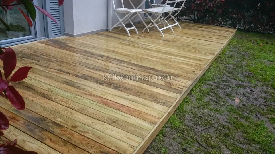 Islak ve çamurlu çimenlik kenarında yeni döşenmiş açık renkli deck; arkada beyaz katlanır sandalyeler