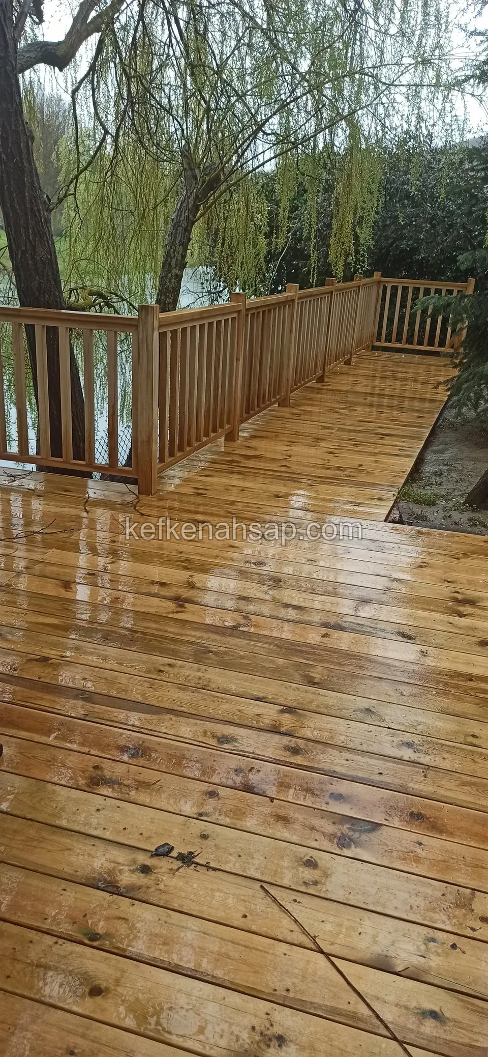Nasse glänzende Kiefern-Deckfläche mit einem Holzgeländer zum See, dahinter eine Weide