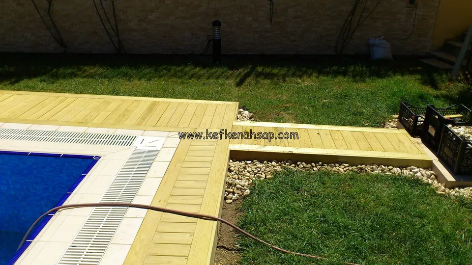 Izgaralı havuz kenarını saran açık renkli deck ve çimeni geçen dar bir deck geçidi; çakıl şeritli kenar