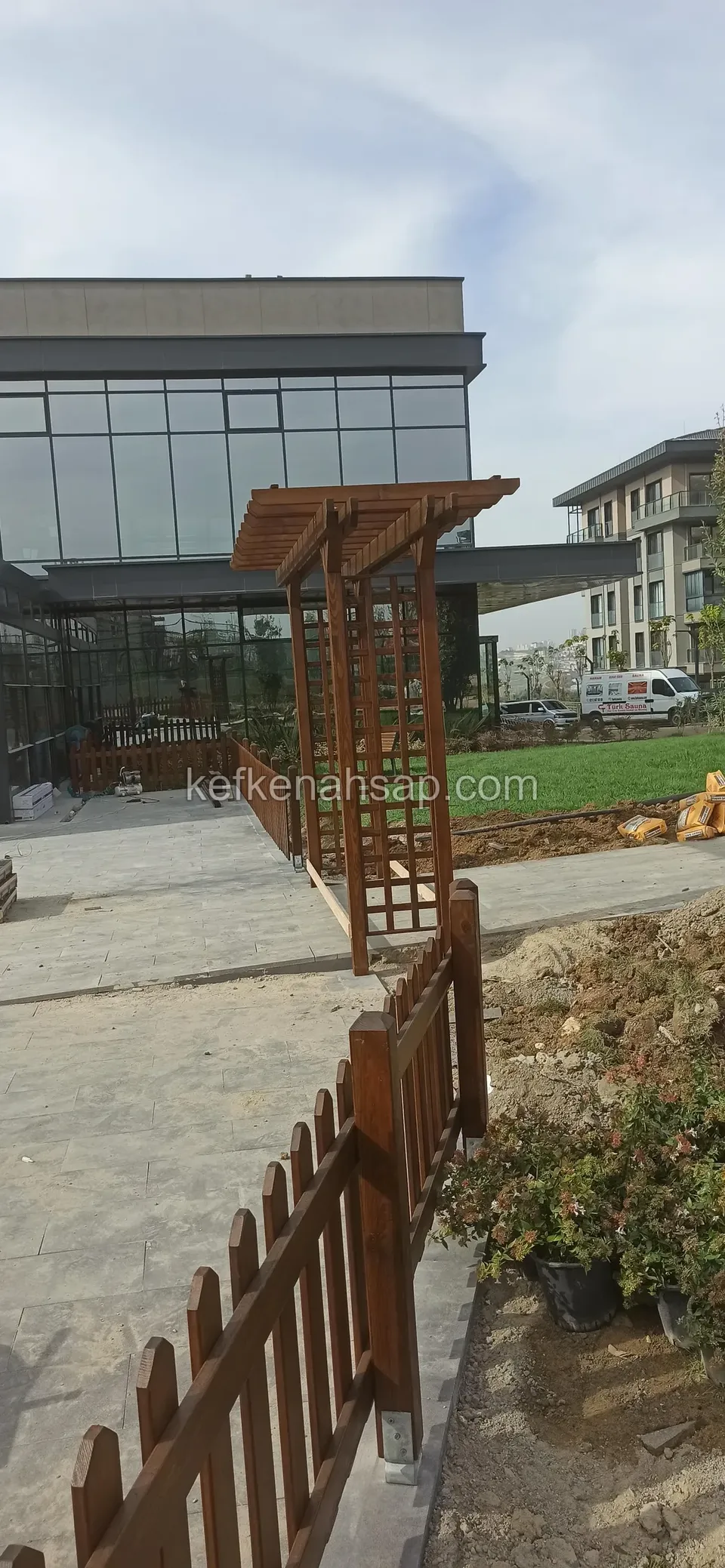 Taş zeminde uzakta kafes pergola kemeri, önde ahşap çıtalı çit ve çalı saksıları, yanında toprak yığını